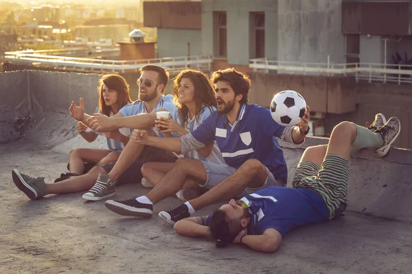 Group of young friends watching a football match on a building rooftop, cheering and drinking beer.