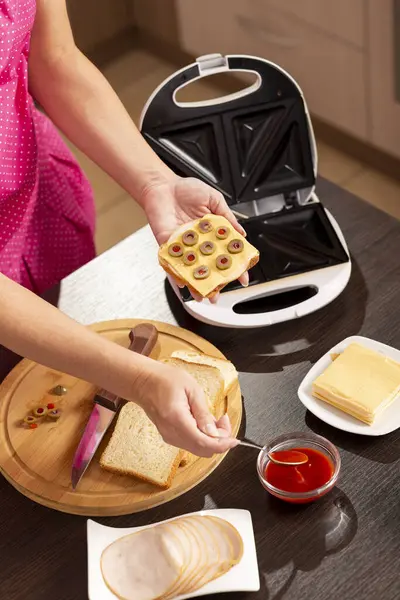 Kadın ellerinin bir sandviçi tuttuğu ve üzerine kaşık ile ketçap koyduğu yüksek açılı manzara; kahvaltıda sandviç makinesinde sıcak sandviç yapan kadın.