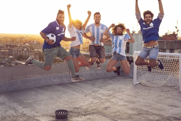 Group of young friends having fun on a building rooftop after a football match; jumping, all caught in the air with the cityscape and urban sunset in the background