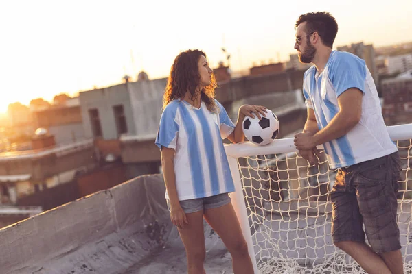 Couple in love wearing football jerseys, standing on a building rooftop after a match and enjoying a beautiful sunset over the city