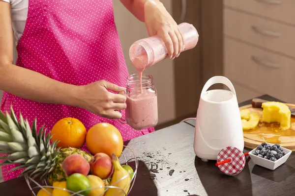 Bir kavanoza yeni yapılmış ahududu smoothie dolduran kadın elleri; mutfakta önlük giymiş bir kadın, kahvaltıda smoothie hazırlıyor.