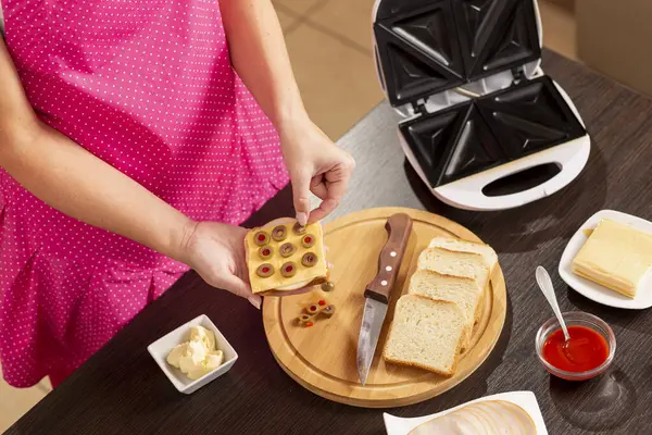 Bir sandviçe zeytin ekleyen kadın elleri, sandviç makinesinde kahvaltıda sıcak sandviç yapan kadın. Sandviçteki zeytinlere odaklan.