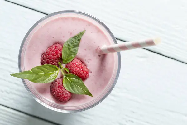 Top view of a raspberry smoothie glass set on a wooden boards, decorated with drinking straw, three raspberries and mint leaves