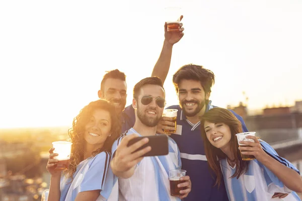 Group of football fans drinking beer and having fun before the game, taking a selfie on a building rooftop terrace. Focus on the girls
