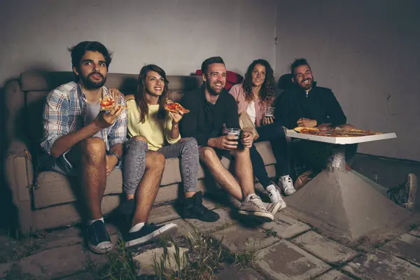 Group of young friends watching a movie on a building rooftop terrace, eating pizza, drinking beer and having fun