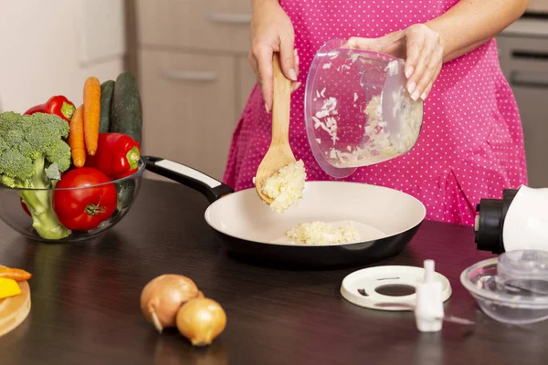 Mutfak tezgahındaki soğan kasesinin mutfak kaşığıyla doğranmış soğanı kızartma tavasına koyan kadın elleri.