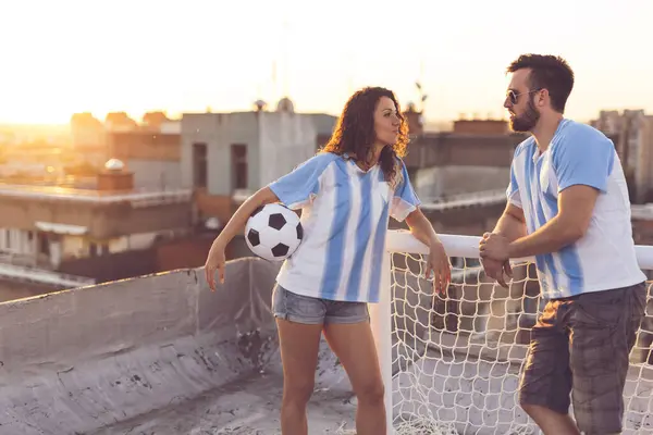 Couple in love wearing football jerseys, standing on a building rooftop after a match and enjoying a beautiful sunset over the city
