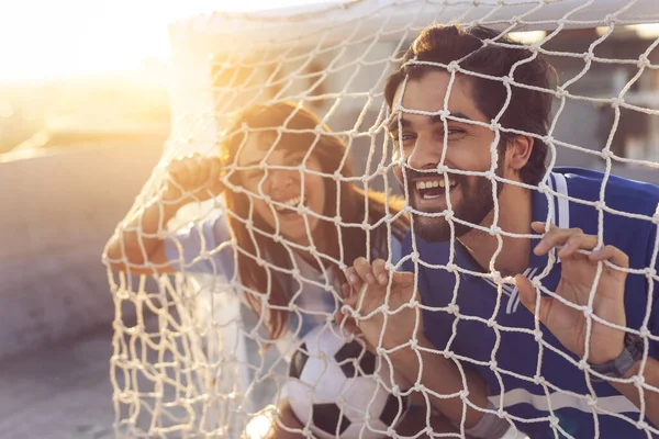 Couple in love wearing football jerseys on a building rooftop after a match, peeking through a goal net and having fun