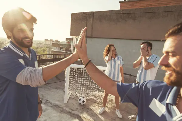 Group of young friends having fun playing football on a building rooftop terrace, winning team celebrating scoring a goal, doing high five while losers mourn failure