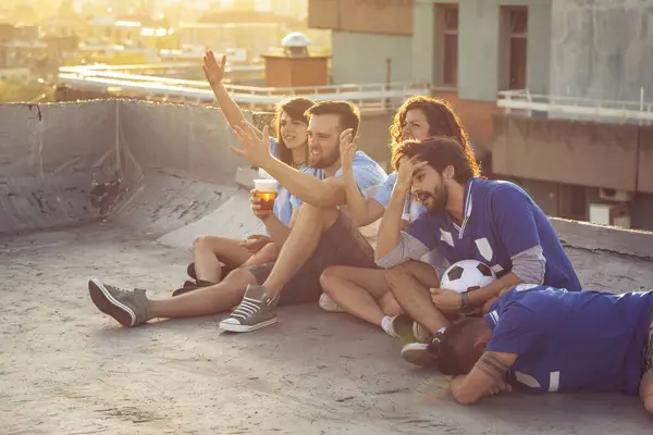 Group of young friends watching a football match on a building rooftop, cheering and drinking beer. Focus on the couple in the middle