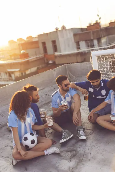 Group of young people sitting on a building rooftop, wearing jerseys, taking a break from a football match, enjoying sunset over the city