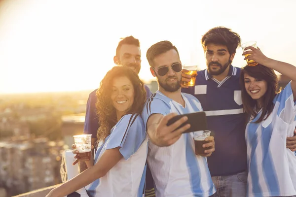 Group of football fans drinking beer and having fun before the game, taking a selfie on a building rooftop terrace. Focus on the girl on the left