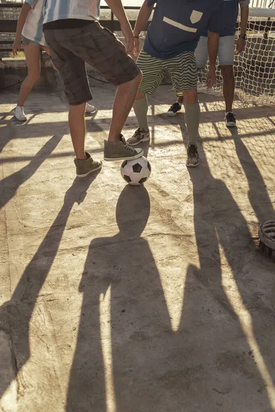 Group of young friends wearing jerseys playing football on a building rooftop terrace on a sunny summer day, detail of legs kicking the ball