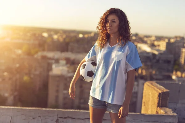 Young woman wearing a football jersey standing on a building rooftop, holding a ball and watching a sunset over the city.