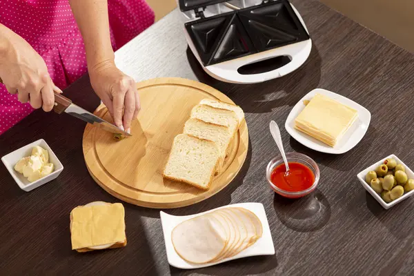 Kadın ellerinin kesme tahtasındaki sandviçler için zeytin keserken yüksek açılı görüntüsü kahvaltıda sandviç makinesinde sıcak sandviç yapan kadın.