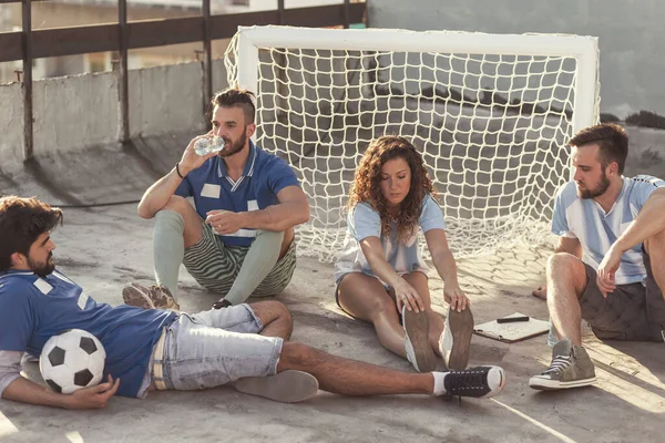 Group of young friends relaxing on a building rooftop terrace after playing a football match, sitting by the goal, drinking water and hanging out