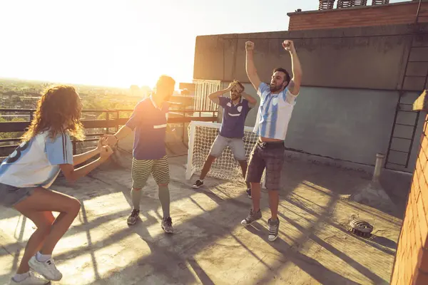 Group of young friends having fun playing football on a building rooftop. One team scored a goal celebrating while the other one anxious because of losing the game