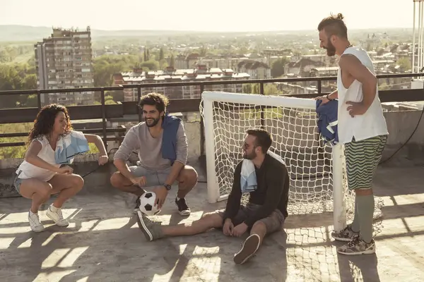 Group of young people relaxing after playing football on building rooftop terrace, having fun spending leisure time togehter outdoor
