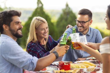 Bir grup genç arkadaş dışarıda barbekü yapıyorlar, masada otururken kadeh kaldırıyorlar. Ortadaki kıza odaklan.