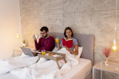 Güzel, genç bir çift pijama giyer, yatakta kahvaltı yaparken dizüstü bilgisayar kullanarak internette gezinir ve eğlenir.