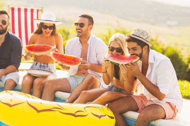 Havuz başında bir yaz partisinde bir grup arkadaş, yüzme havuzunun kenarında oturmuş, soğuk karpuz dilimleri yiyor ve eğleniyorlar.