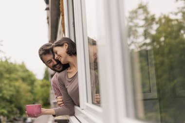 Aşık bir çift açık bir pencereye yaslanıyor, dışarı bakıyor, kahve içiyor ve evde dinleniyor.
