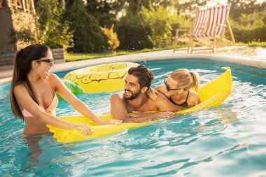 Bir grup arkadaş havuz başında bir yaz partisinde, yüzme havuzunda eğleniyorlar, yüzen yatakta güneşleniyorlar.