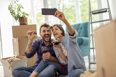 Genç çift yeni bir daireye taşınıyor, şarap içiyor ve ev anahtarlarıyla selfie çekerken eğleniyor.