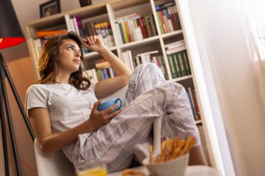 Pijama giyen güzel genç bir kadın, oturma odasının penceresinin yanında oturuyor, elinde bir fincan kahve tutuyor ve anın tadını çıkarıyor.