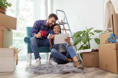 Birbirine aşık bir çift, tablet bilgisayar ve dergiler kullanarak ev dekorasyonu fikirleri ararken kahve içip eğleniyor.