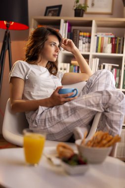 Pijama giyen, oturma odasının penceresinin yanında oturan ve sabah kahvesinin tadını çıkaran genç ve güzel bir kadın.