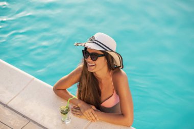 Bikini, güneş gözlüğü ve şapka takan çekici genç bir kadın havuzda dikiliyor, kenara yaslanıyor, kokteyller içiyor ve güneşleniyor.