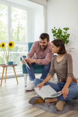 Güzel bir çift evde dinleniyor, bir adam koltukta oturuyor, tablet bilgisayar kullanıyor ve kadın yerde oturup dergi okuyor.