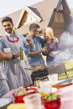 Bir grup arkadaş arka bahçede barbekü yapıyor, bira içiyor ve güneşli bir yaz gününde eğleniyorlar.