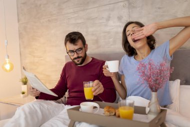 Genç çift pijama giyiyor, sabahları yatakta oturuyor, evde dinleniyor kadın esniyor ve kahve içiyor erkek gazete okurken.