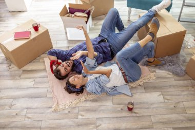 Birbirine aşık çiftlerin yeni evlerinin zemininde yatıp tablet bilgisayar kullanarak öpüşmelerini gösteren yüksek açılı görüntüler.