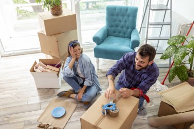 Karton kutulara bir şeyler paketleyen, taşınmaya hazırlanan bir çiftin yüksek açılı görüntüsü. Kadın kırılgan şeyleri kağıda sararken, erkek kutuları paket makinesiyle bantlıyor.