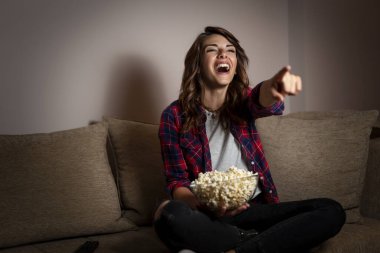 Genç ve güzel bir kadın oturma odasındaki kanepede oturuyor, TV 'de komedi izlerken eğleniyor ve patlamış mısır yiyor.