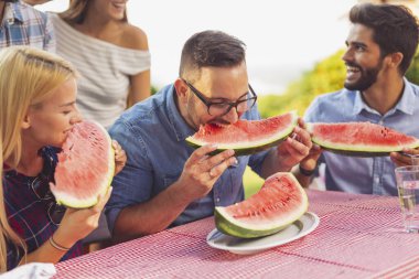 Bir grup arkadaş dışarıda öğle yemeği yiyor, taze karpuz dilimleri yiyor ve eğleniyorlar.