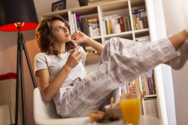 Pijama giyen güzel genç bir kadın, oturma odasının penceresinin yanında oturuyor, elinde bir fincan kahve tutuyor ve anın tadını çıkarıyor.