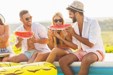 Havuz başında bir yaz partisinde bir grup arkadaş, yüzme havuzunun kenarında oturmuş, soğuk karpuz dilimleri yiyor ve eğleniyorlar.