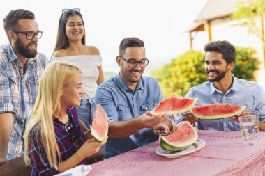 Bir grup arkadaş dışarıda öğle yemeği yiyor, taze karpuz dilimleri yiyor ve eğleniyorlar.
