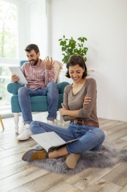Güzel bir çift evde dinleniyor, bir adam koltukta oturuyor, tablet bilgisayar kullanıyor ve kadın yerde oturup dergi okuyor.