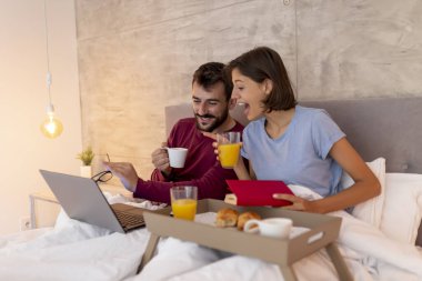 Güzel, genç bir çift pijama giyer, yatakta kahvaltı yaparken dizüstü bilgisayar kullanarak internette gezinir ve eğlenir.