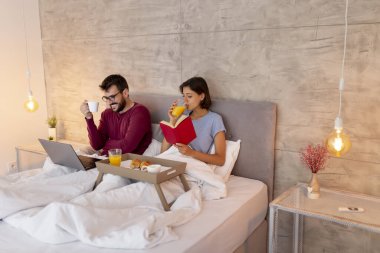 Güzel, genç aşık bir çift pijama giyiyor, kadın kitap okuyor ve erkek yatakta kahvaltı ederken dizüstü bilgisayarla internette sörf yapıyor.