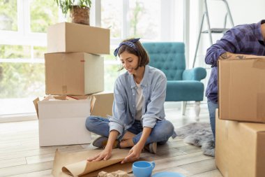 Güzel genç bir kadın yerde oturuyor, kırılgan şeyleri kağıda sarıp karton kutulara dolduruyor, taşınmaya hazırlanıyor.