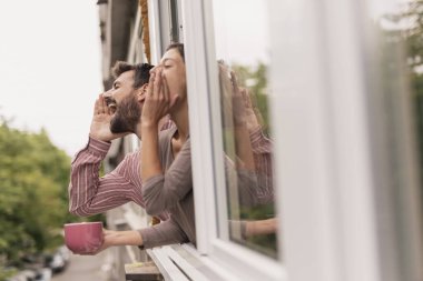 Güzel genç bir çift açık bir apartman penceresinden eğilmiş, el sallıyor ve bağırıyor, yoldan geçen arkadaşlarını arıyor.