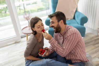 Birbirine aşık çiftler yeni evlerinde yerde oturmuş çilek yiyor, gülüyor ve eğleniyorlar.