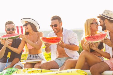 Havuz başında bir yaz partisinde bir grup arkadaş, yüzme havuzunun kenarında oturmuş, soğuk karpuz dilimleri yiyor ve eğleniyorlar.