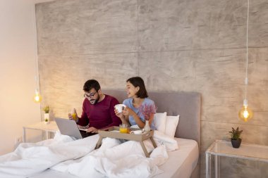 Aşık bir çift yatakta uzanıyor, hafta sonu sabahının tadını çıkarıyor, kahvaltı yapıyor ve internette dizüstü bilgisayarla sörf yapıyor.